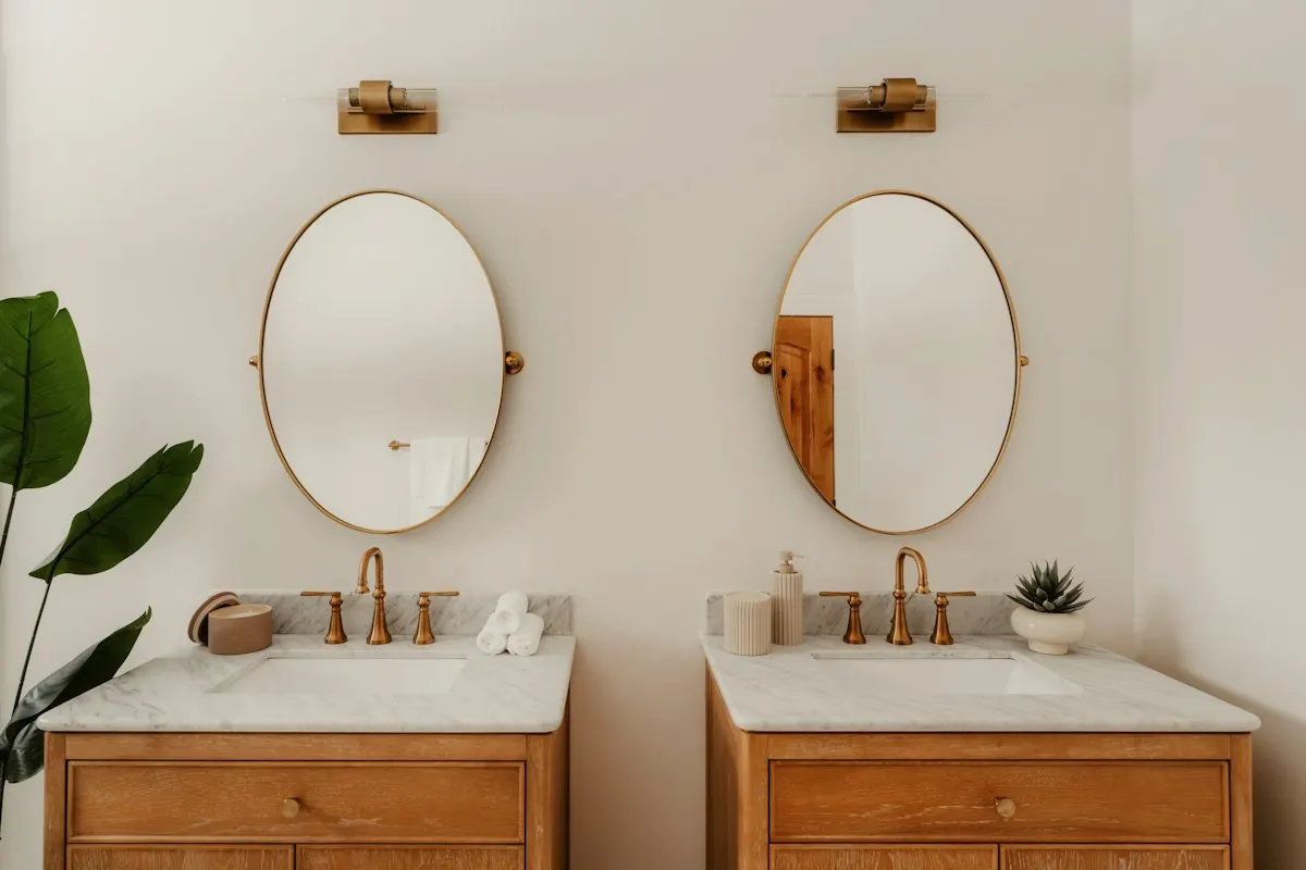 Twin timber vanities with oval mirrors, brass tapware and leafy accents in a bright coastal bathroom